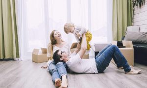 family of three laying on the floor in a house surrounded by brown boxes and a big window in the background